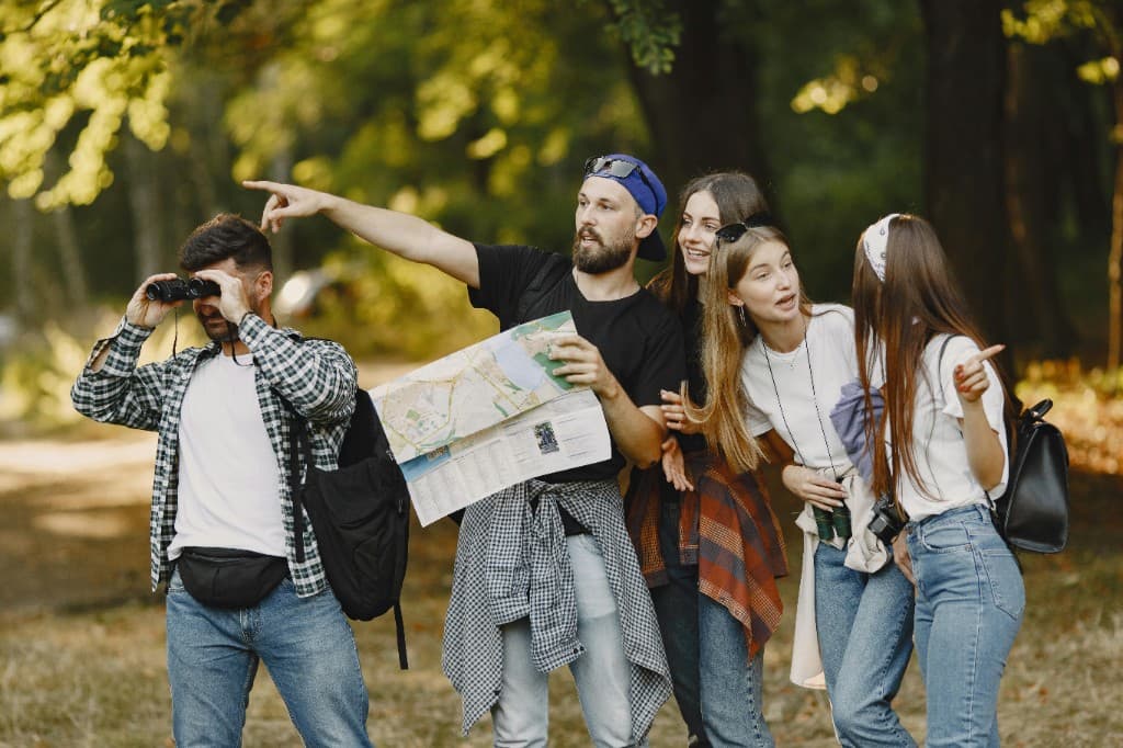 Group of friends exploring outdoors with a map, binoculars, backpacks, and cameras during a scavenger hunt style adventure.
