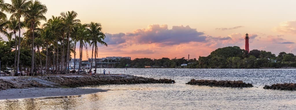 Jupiter Inlet Lighthouse at sunset overlooking the water with palm trees along the Florida shoreline.