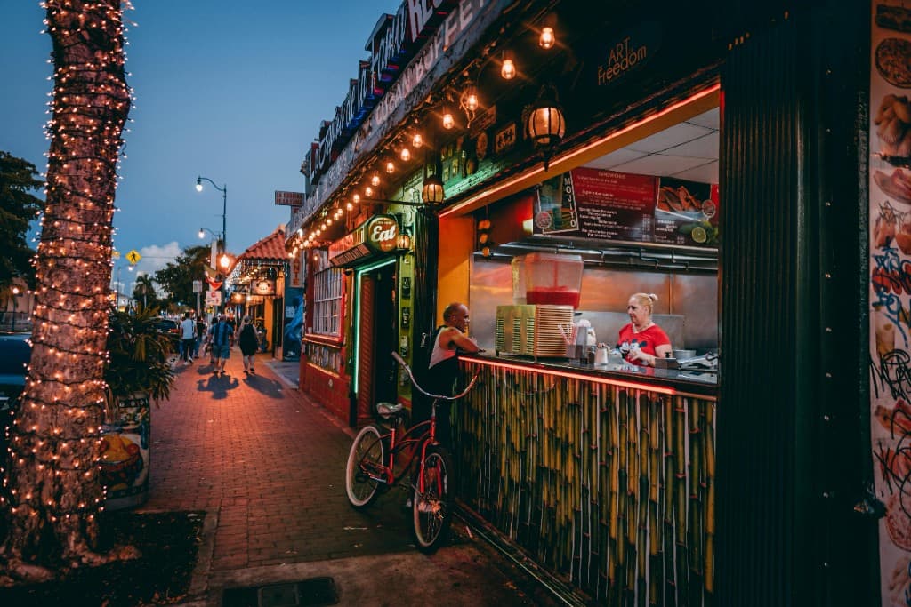 Evening street scene in Miami’s Little Havana with a walk-up food window, string lights, pedestrians, and a bicycle.