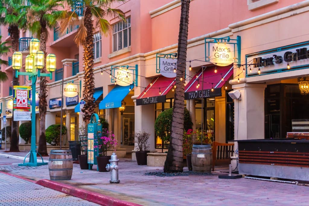 Palm lined restaurant row at Mizner Park in Boca Raton, Florida with colorful storefronts, string lights, and outdoor dining areas.