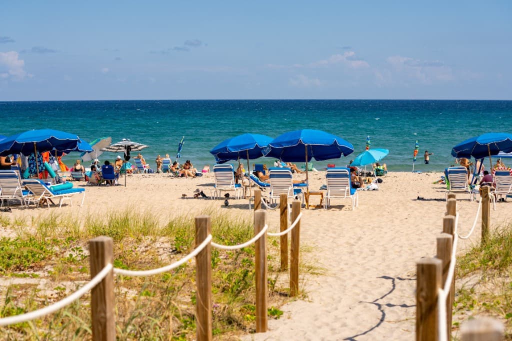 Beach umbrellas and lounge chairs on Delray Beach with ocean waves, sand dunes, and a sunny blue sky.