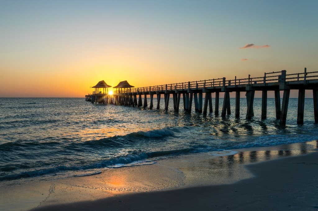 Naples beach pier at sunset with gentle waves, golden sky, and the shoreline reflecting the evening light.