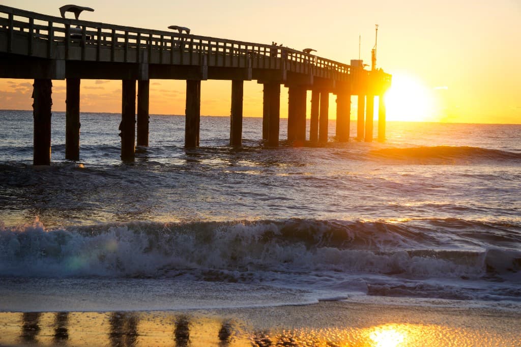 St. Augustine Beach pier at sunrise with ocean waves, golden sky, and sunlight reflecting on the shoreline.